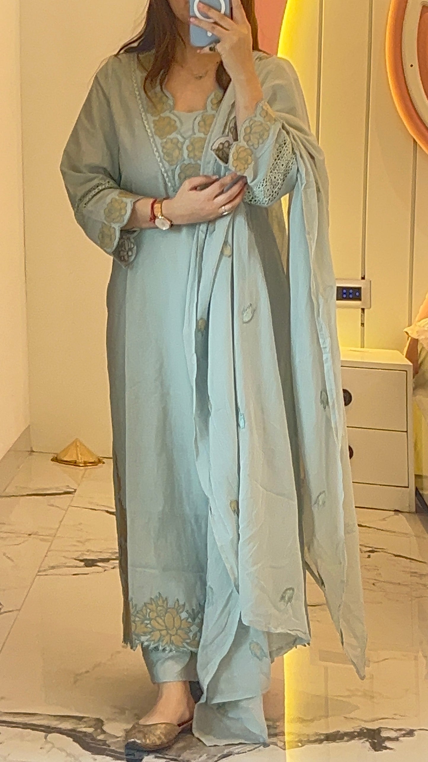 Person wearing a light blue embroidered outfit in a room with a mirror and white dresser.